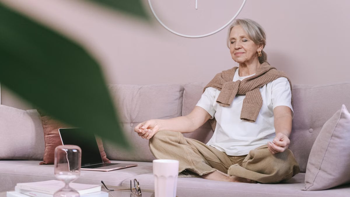 Elderly woman sitting on a sofa meditating with eyes closed, in a peaceful home setting.